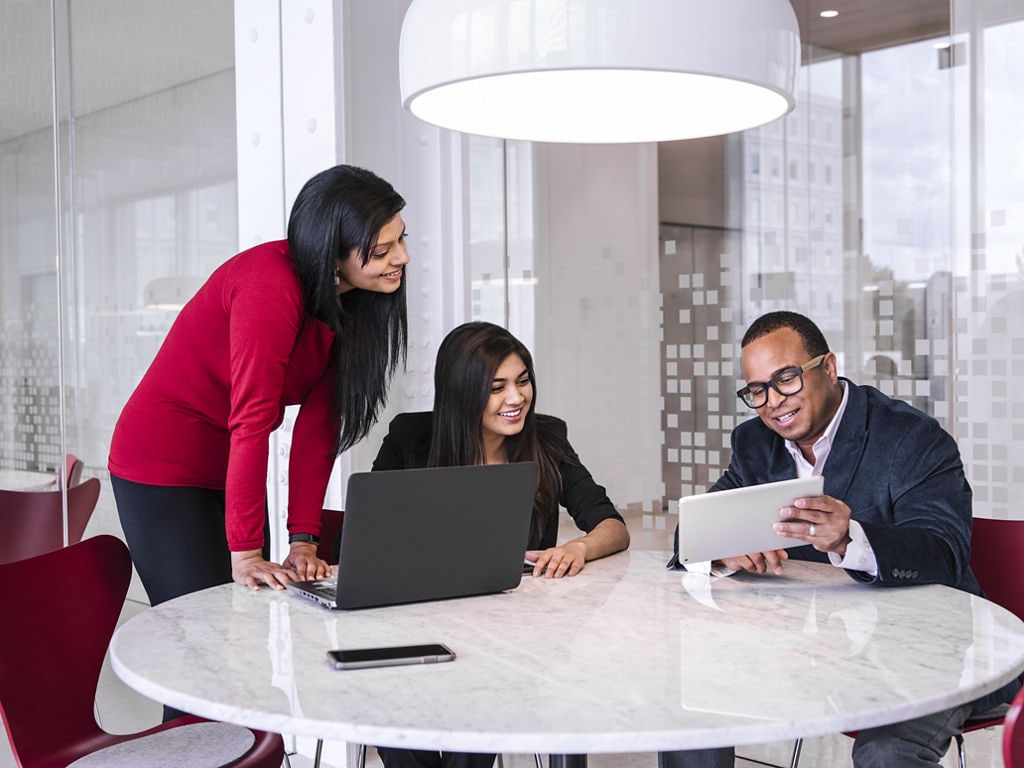 Three professionals collaborate around a round table in a modern office setting, with one person using a laptop and another holding a tablet.
