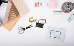Top down view of a home office or office desk. On the desk are common stationery items like Scotch Double-Sided Tape, Scotch™ Brand Scissors. The featured action depicts the person creating labels for a small mailing envelope.