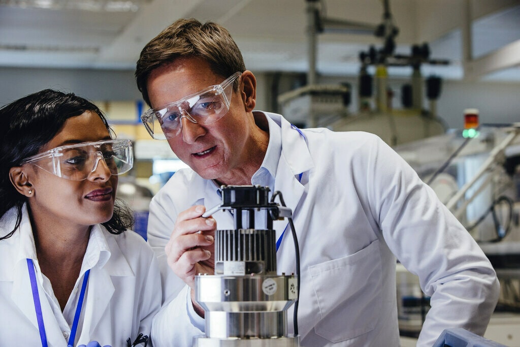 Two individuals wearing white lab coats are closely examining a piece of laboratory equipment in a research or industrial setting.