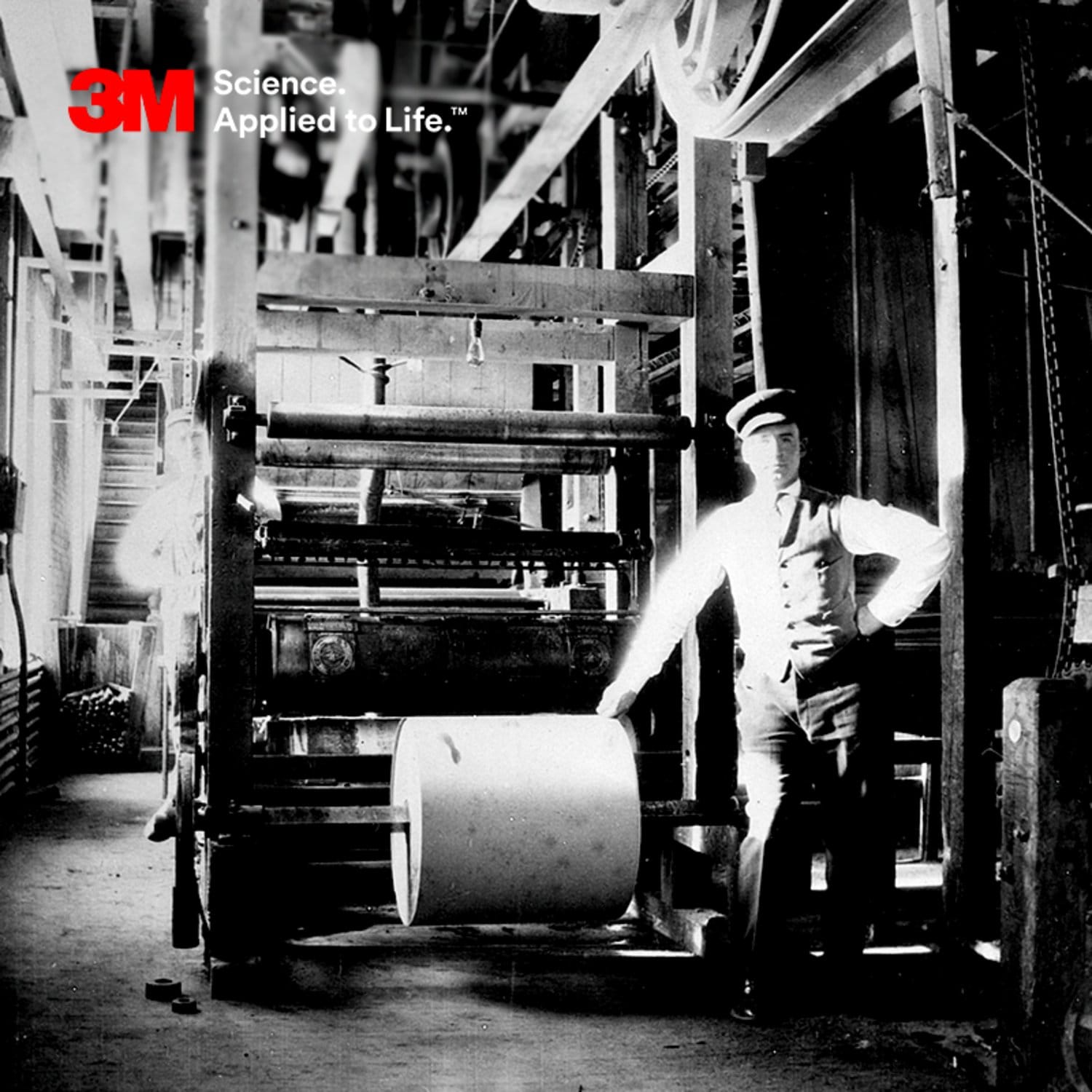 A historical black and white scene featuring a person in early 20th century attire standing next to industrial machinery.