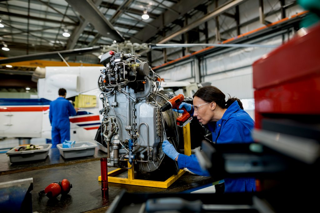 A woman with a ponytail and safety glasses works on a helicopter propeller engine.