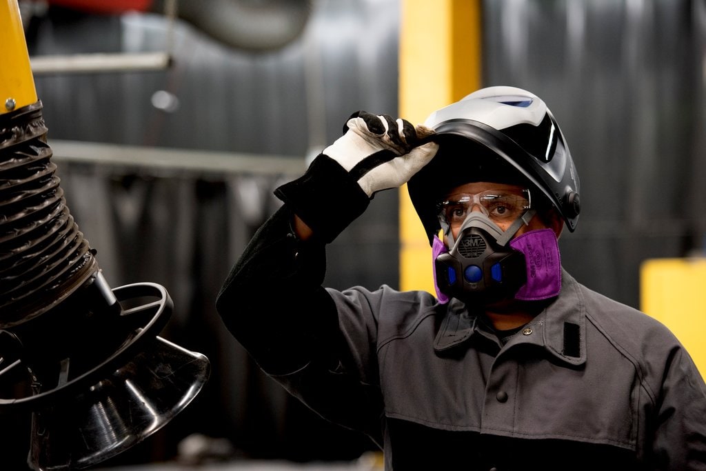 Worker in industrial protective gear adjusts a welding helmet while wearing gloves and a respirator in a manufacturing setting.