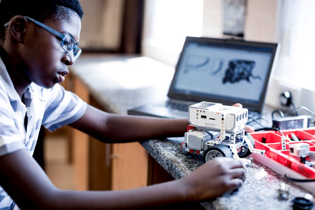 A person is working on assembling a small robot on a countertop, with a laptop displaying technical diagrams in the background.