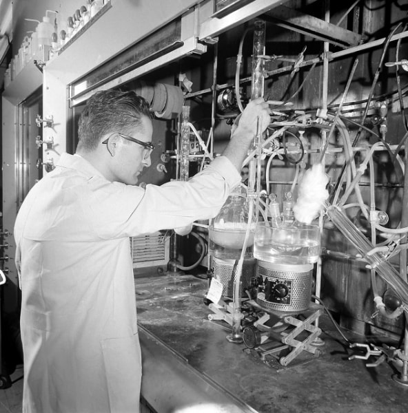 A scientist or technician in a lab coat is working with a complex setup of glassware and tubing, likely conducting an experiment or chemical process.