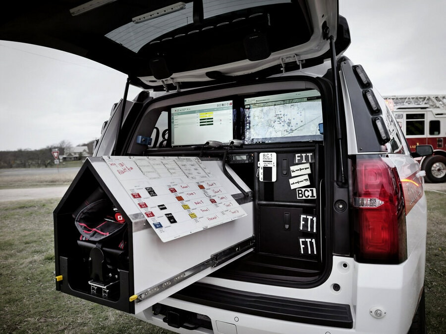 Mobile command workstation set up in the rear of a white vehicle, featuring a fold-out control panel, computer monitor, keyboard, and radio equipment.