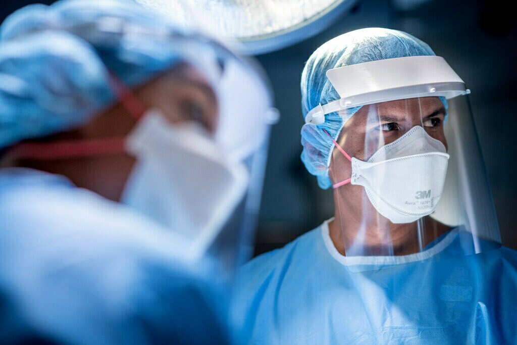 Two healthcare professionals look to their left wearing surgical gowns, face shields and N95 masks.