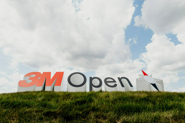 The 3M Open sign is displayed on a grassy hill, with a blue sky and clouds in the background.