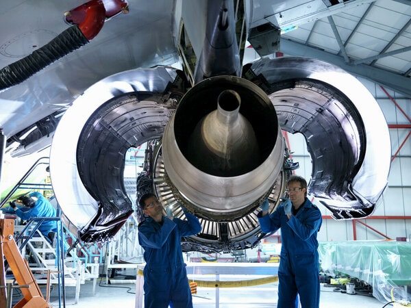 Two technicians in blue uniforms inspect the underside of an aircraft engine in a hangar.