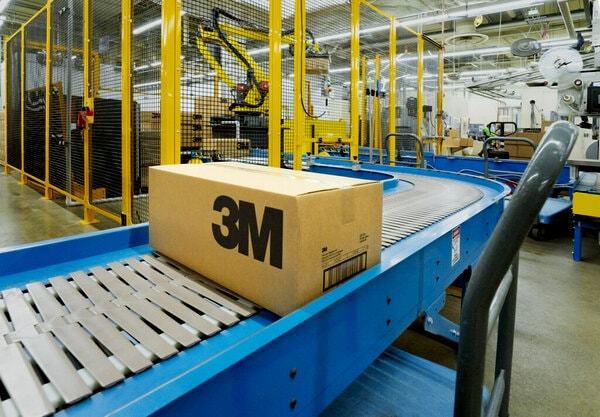 A cardboard box with the 3M logo is placed on a blue conveyor belt in an industrial setting.