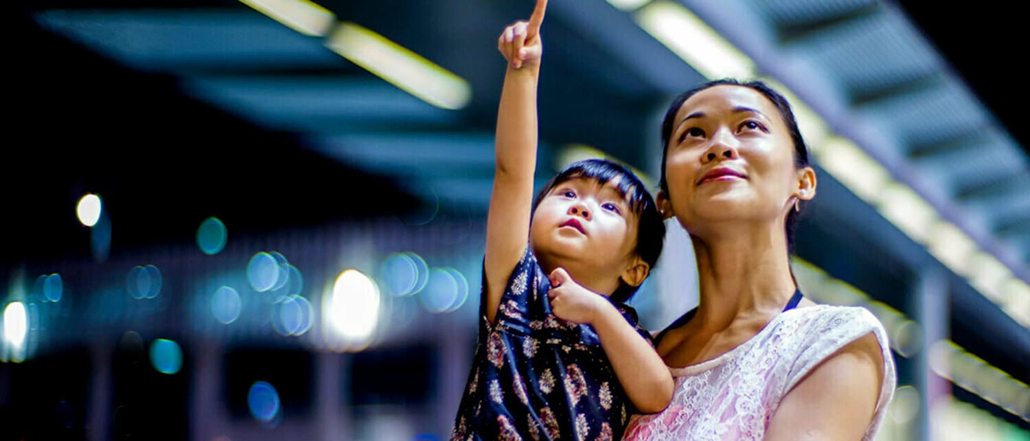 A woman holding a child in her arms while both are gazing upwards with the child pointing upwards against a pixelated background of a mass transit train platform.