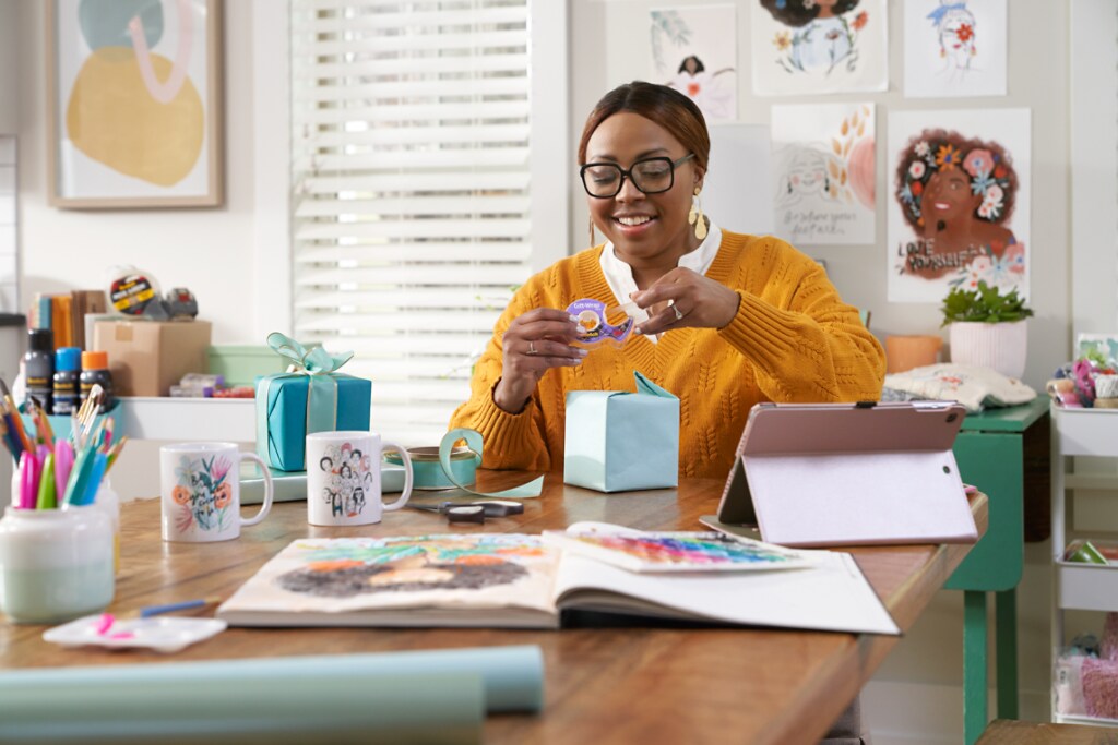 A young black woman wearing thick rimmed glasses and a yellow sweater sits at a kitchen table wrapping a small purple present.