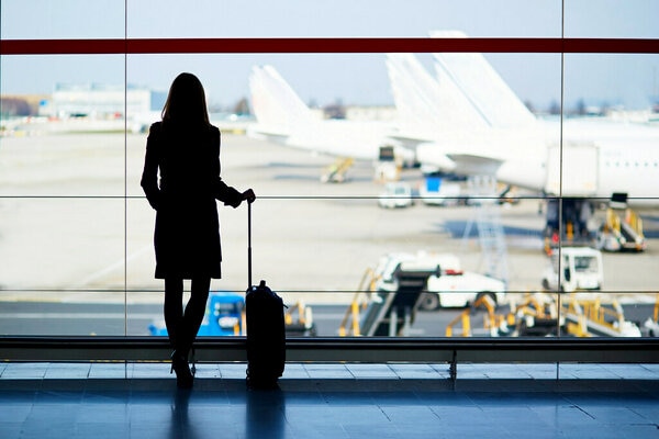 A person with long hair stands indoors, gazing out at an airport runway with airplanes in the background.