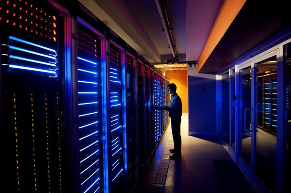 Technician in a dimly lit data center aisle working on server racks, with rows of hardware glowing blue and a warm orange light at the end of the corridor.