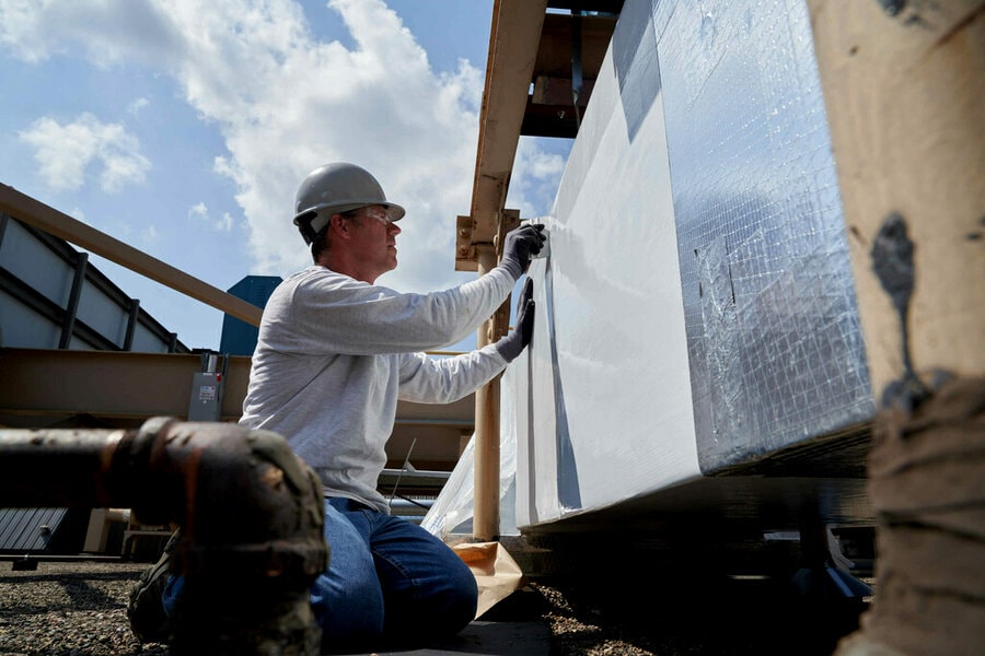 Two construction workers in orange safety gear install 3M weather barrier wrap on the exterior walls of a concrete block building under construction.