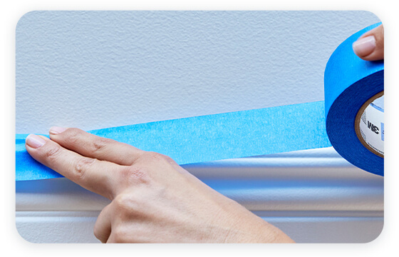 A hand applying a strip of blue painter tape along the edge of a white wall, ensuring a smooth and even application.