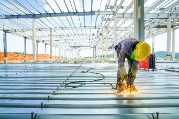 A construction worker wearing a yellow hard hat and protective gear is welding metal beams on a construction site.