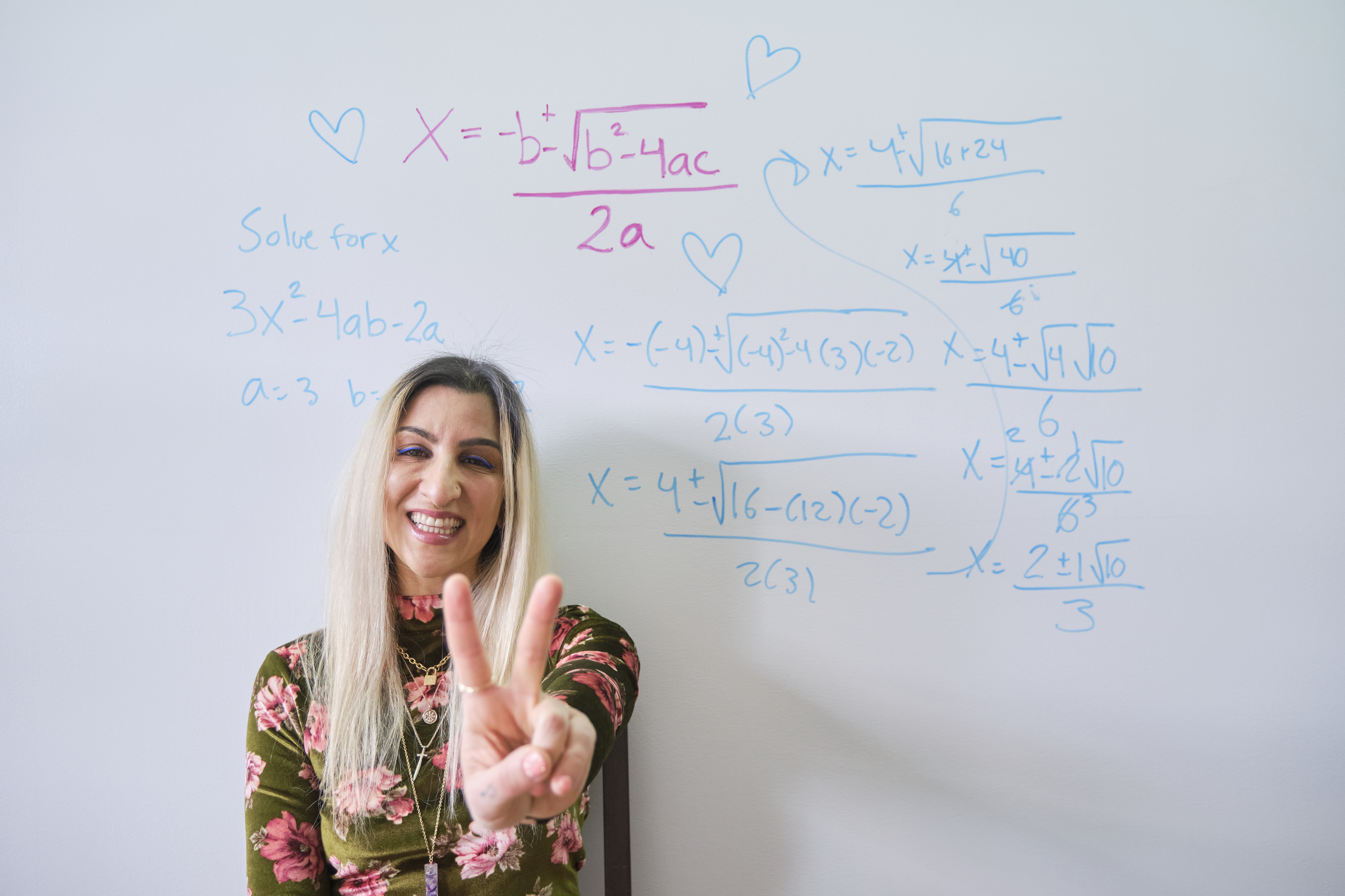 Vanessa Vakharia, a math teacher give a peace sign infront of a white board that contains a math equation.