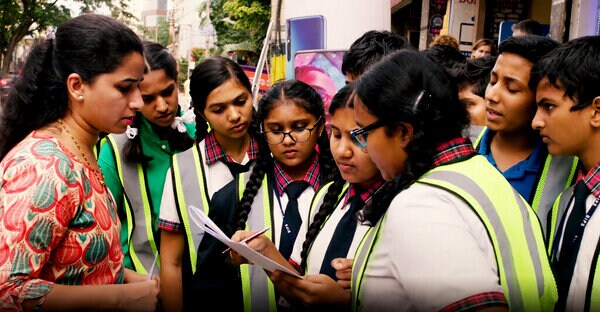 A group of students wearing school uniforms and reflective vests are gathered outdoors, engaging in a discussion with a woman holding a clipboard.