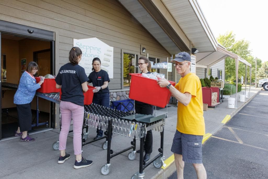 A group of people are working together to move red bins using a conveyor system outside a building labeled Stepping Stones.