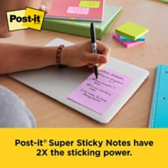 This is an over the shoulder image of a person at a desk with a closed computer and the person is writing on the 4x6 lined Post-it Note.