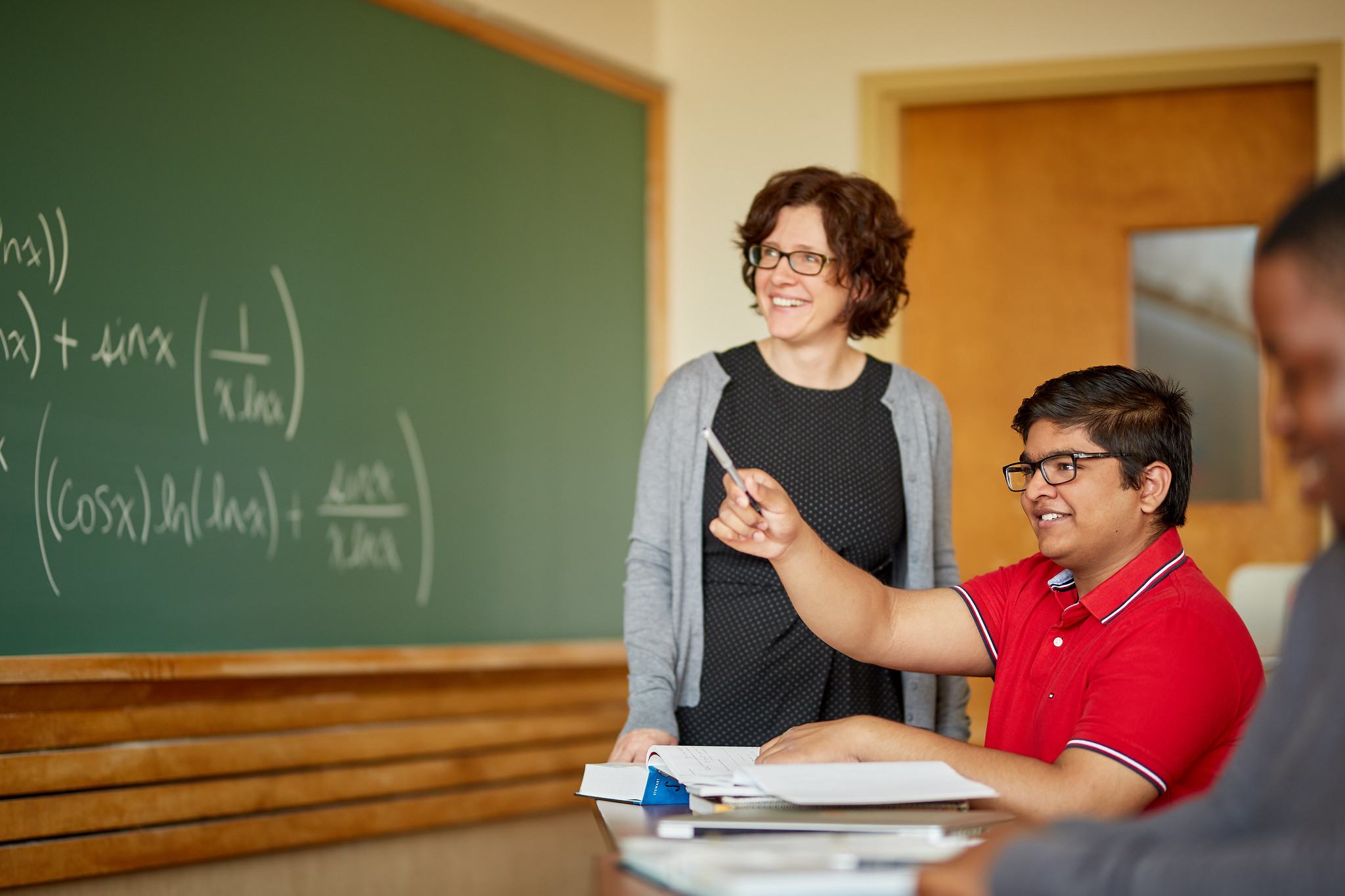 Canadian Educator Anna Stokkes stands beside a sitting student who gestures ahead. She wears a grey cardigan and glasses and behind her is a black board with equations on it. 