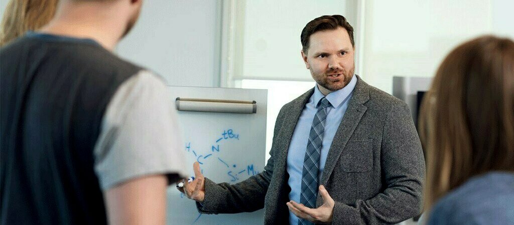 Canadian Educator Brett McCollum wears a grey suit and blue shirt while carrying out a lesson on a white board in front of students who face him. 