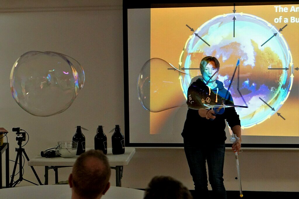 Canadian Educator Joanne O’Meara stands at the front of a dimly lit room creating giant bubbles in front of a screen with a scientific explanation projected on to it. 