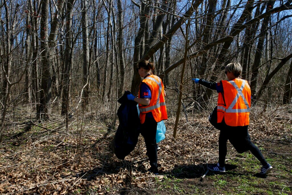 A woman with brown hair in a ponytail leads the way. A woman with short blonde hair walks behind her pointing ahead to an unseen spot in the 3M tartan forest. Both 3Mers are wearing blue 3M volunteer shirts, orange high visibility vests, blue gloves, and hold garbage bags.  