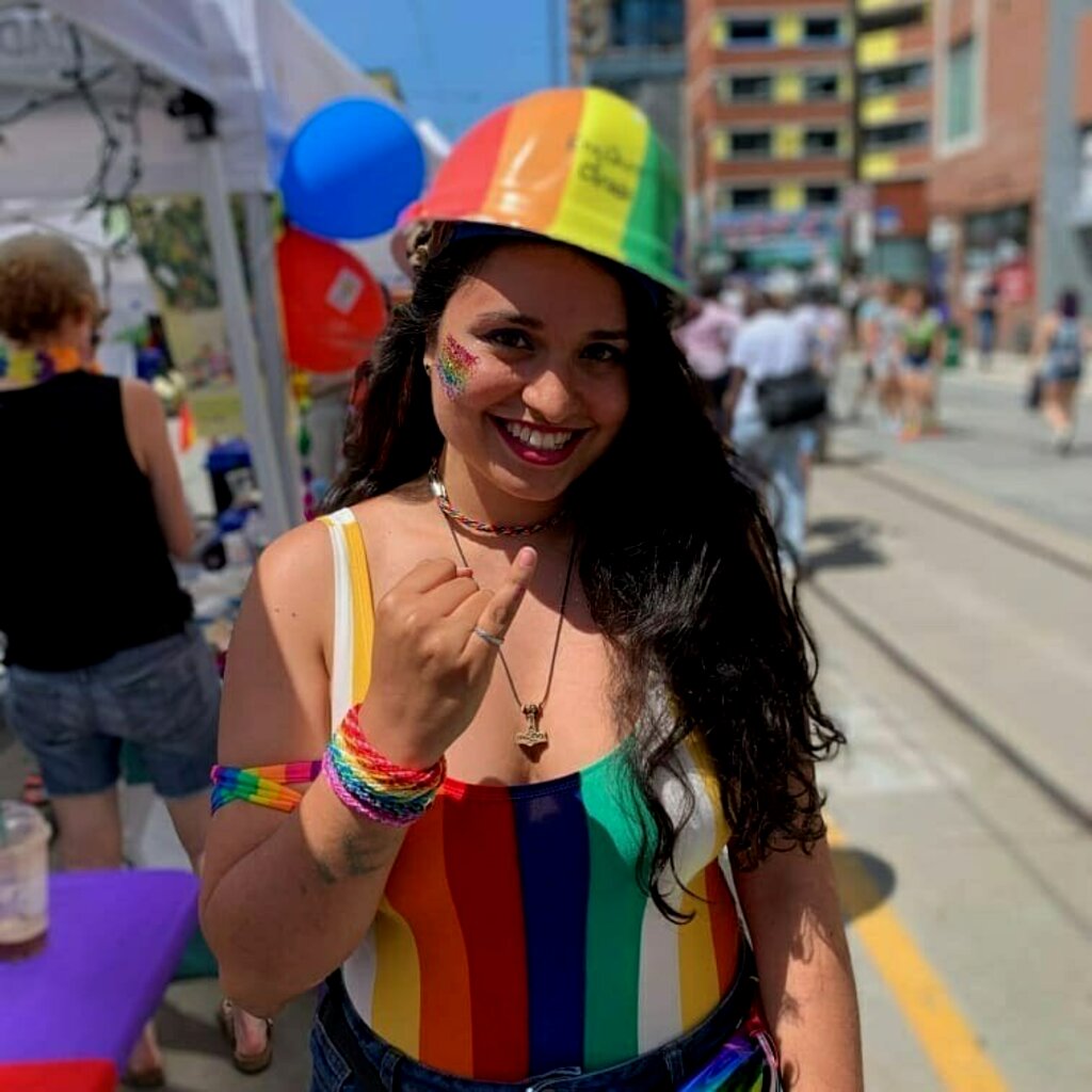 Woman with dark hair smiling and wearing a rainbow coloured construction hat, jumpsuit and armbands holding up her pinky finger.