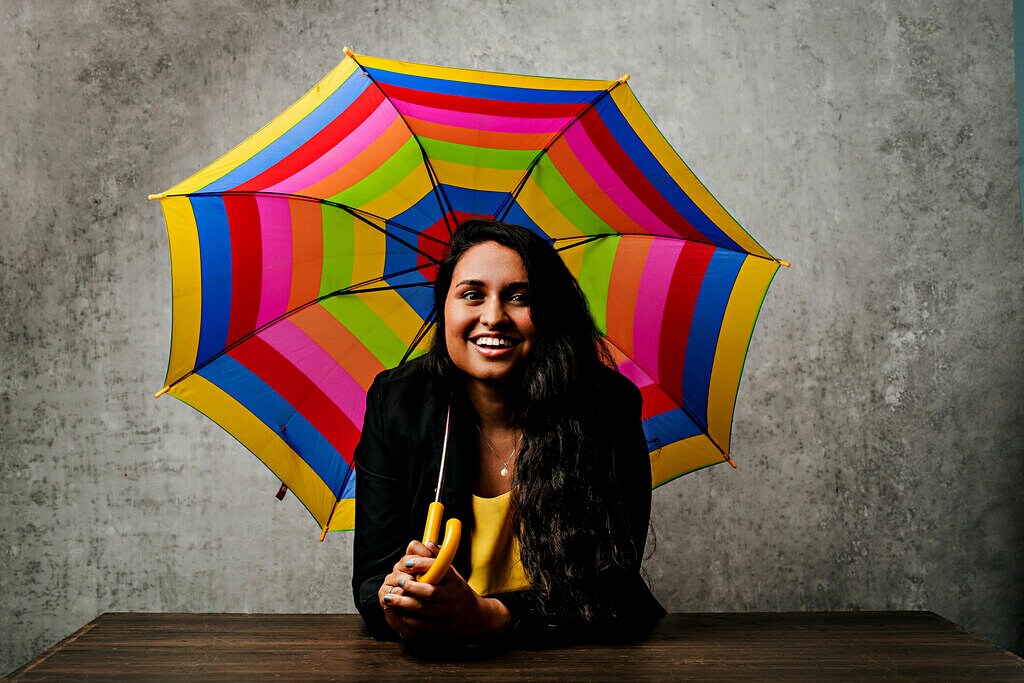 A woman with dark hair and a yellow shirt smiling and holding a rainbow coloured umbrella.