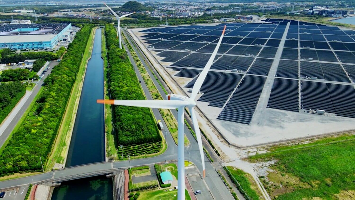 A large wind turbine stands adjacent to a canal, surrounded by lush greenery and in the background, rows of solar panels.
