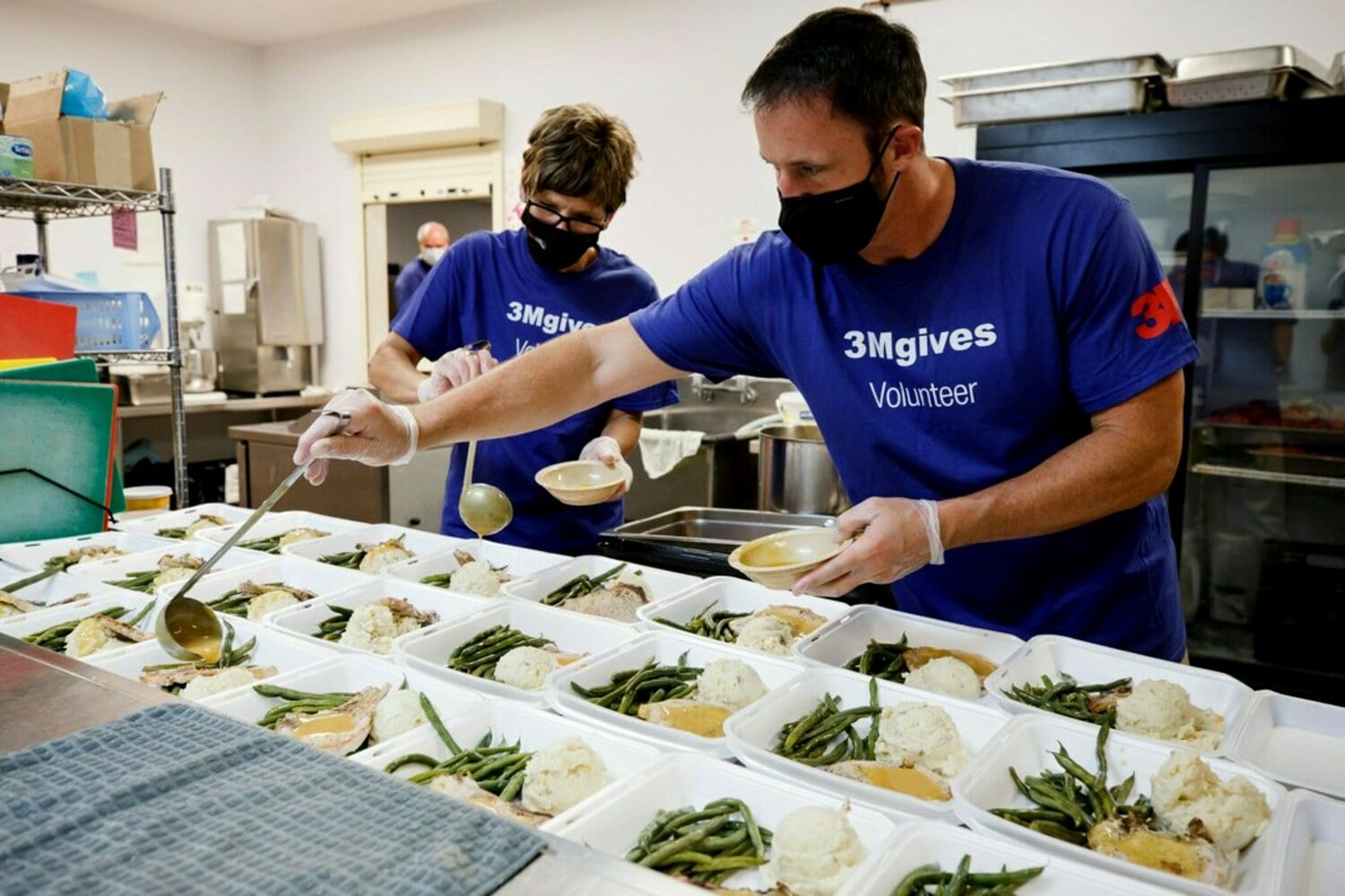 Two individuals wearing blue 3Mgives Volunteer shirts are engaged in a community service activity in a kitchen setting.