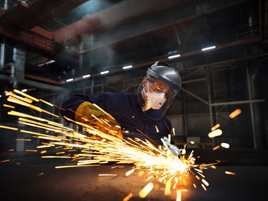 Industrial worker grinding metal in a factory while wearing a face shield, gloves, and protective clothing, with bright sparks flying across the foreground.