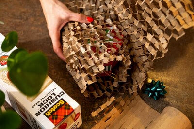 A hand with red nail polish wrapping a gift in brown honeycomb-patterned paper, with a roll of Scotch Cushion Lock protective wrap and a small blue gift bow nearby on a table.