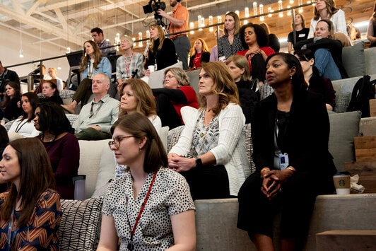 A diverse group of people is seated in a tiered auditorium, attentively listening to a presentation.