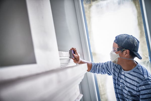 Worker in a striped shirt uses a sanding block to smooth white interior trim or a mantel near a window, wearing a cap and dust mask during home renovation.