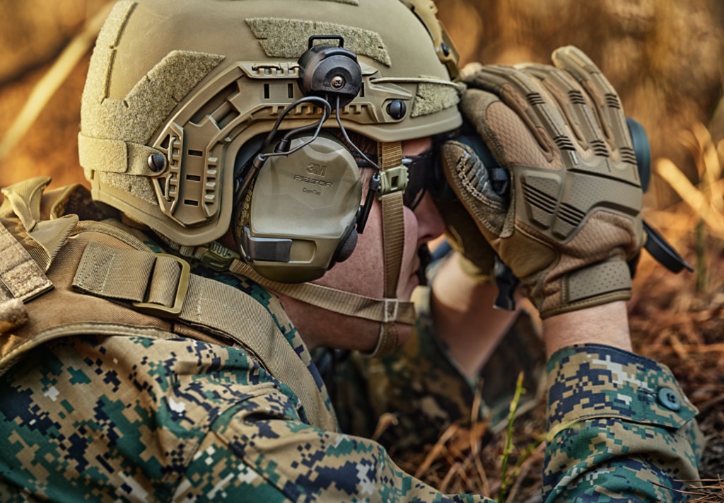 Military soldier wearing a helmet with headset, observing through binoculars.