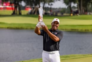 A golfer wearing a black shirt and white pants holds up a clear trophy triumphantly on a golf course, with a water hazard and trees in the background.