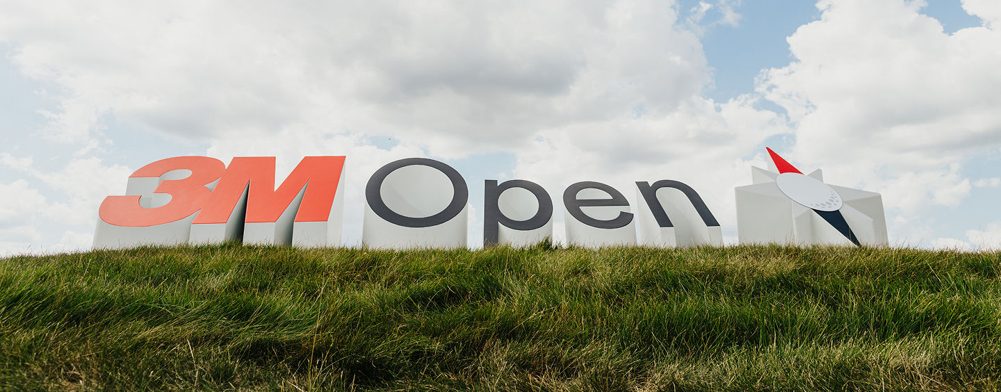 Large 3M Open sign on a grassy hill, set against a partly cloudy sky, indicating the 3M Open golf tournament.