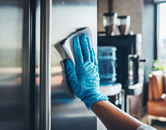 A person wearing blue gloves cleans the stainless steel handle of a refrigerator in an office kitchen area.
