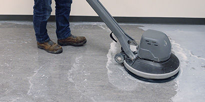 An individual using a handheld floor scrubber on a concrete floor.