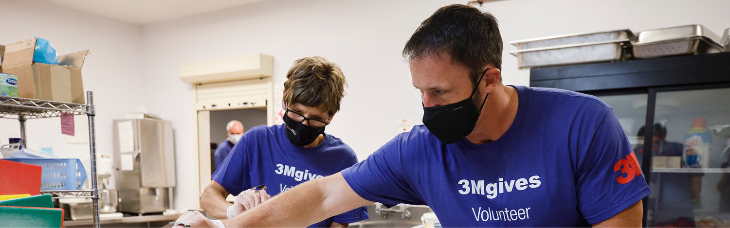 Two individuals wearing blue 3Mgives Volunteer shirts are engaged in a community service activity in a kitchen setting.