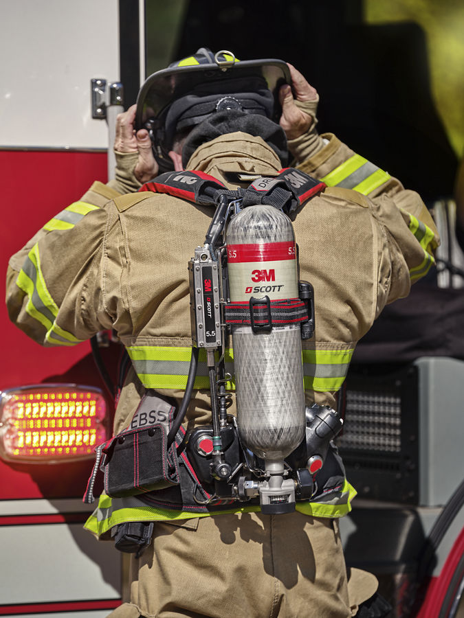 Firefighter in turnout gear wearing a 3M Scott SCBA air cylinder on their back, shown from behind.