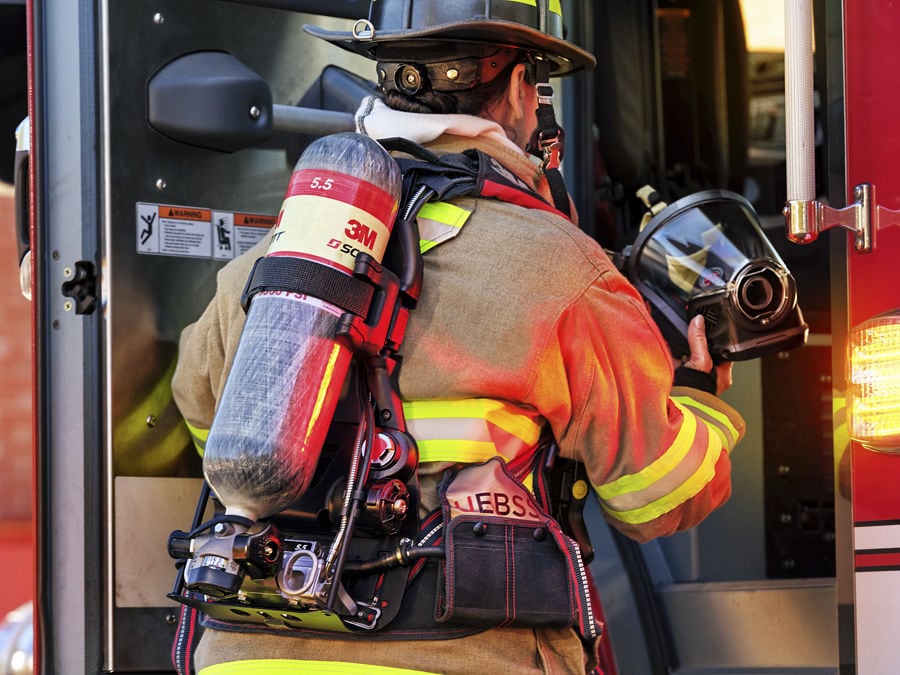 Firefighter in turnout gear enters a fire truck, carrying a helmet and wearing a 3M SCBA air tank.