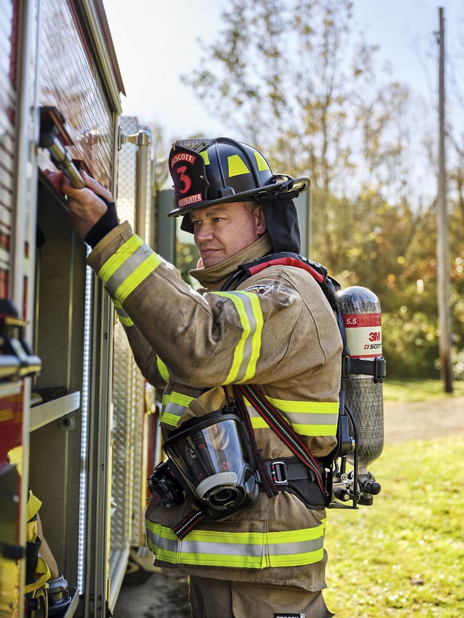 Firefighter in turnout gear stands at the side of a fire truck, reaching toward equipment while wearing a 3M SCBA air tank.