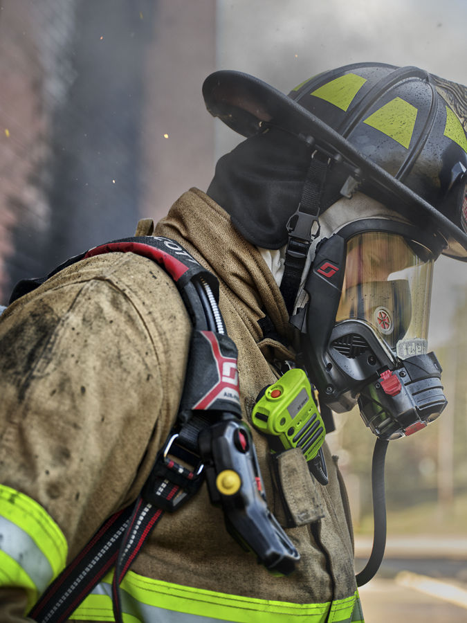 Firefighter in turnout gear wearing a full-face respirator and helmet, with a shoulder radio visible against a smoky background.