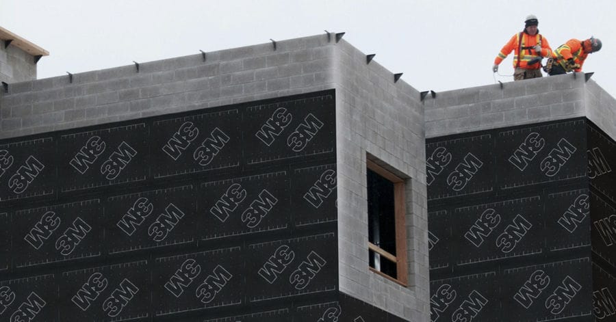 A construction worker in a hard hat and safety glasses applies 3M VentureClad insulation jacketing to HVAC equipment on a rooftop.