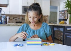 Photograph of a young girl smiling while sitting at a white table in a bright kitchen, using a multicolored stack of sticky notes with several small rolls of decorative tape placed around it.