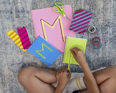 Photograph of overhead view of a child decorating colorful notebooks and paper with glitter tape, including a pink sheet and blue notebook with a large decorative letter “M.”