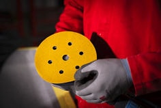 Close-up de um trabalhador usando uma jaqueta de proteção vermelha e luvas de segurança cinza segurando um disco de lixamento circular amarelo acoplado a uma lixadeira elétrica. O disco de lixamento possui múltiplos furos para extração de pó e está posicionado em destaque no primeiro plano. O fundo é escuro e desfocado, enfatizando a ferramenta e o disco abrasivo.